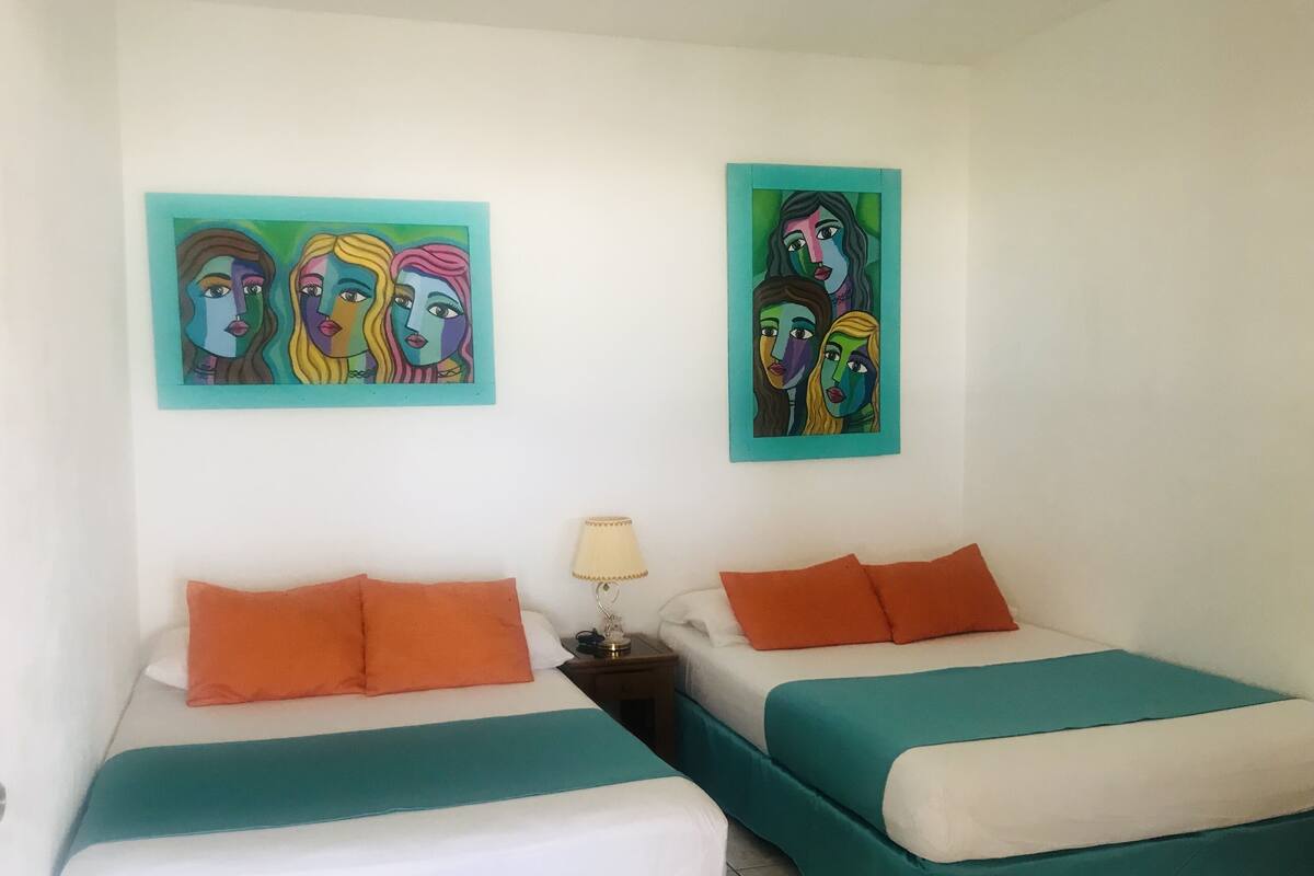 Een hotelkamer met twee bedden, elk met koper- en wit beddengoed, oranje kussens en twee kleurrijke schilderijen aan de muur.