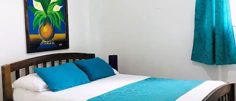 A single bed with a wooden frame, white bedding, and a teal patterned throw. A framed painting of flowers hangs on the wall above the bed.