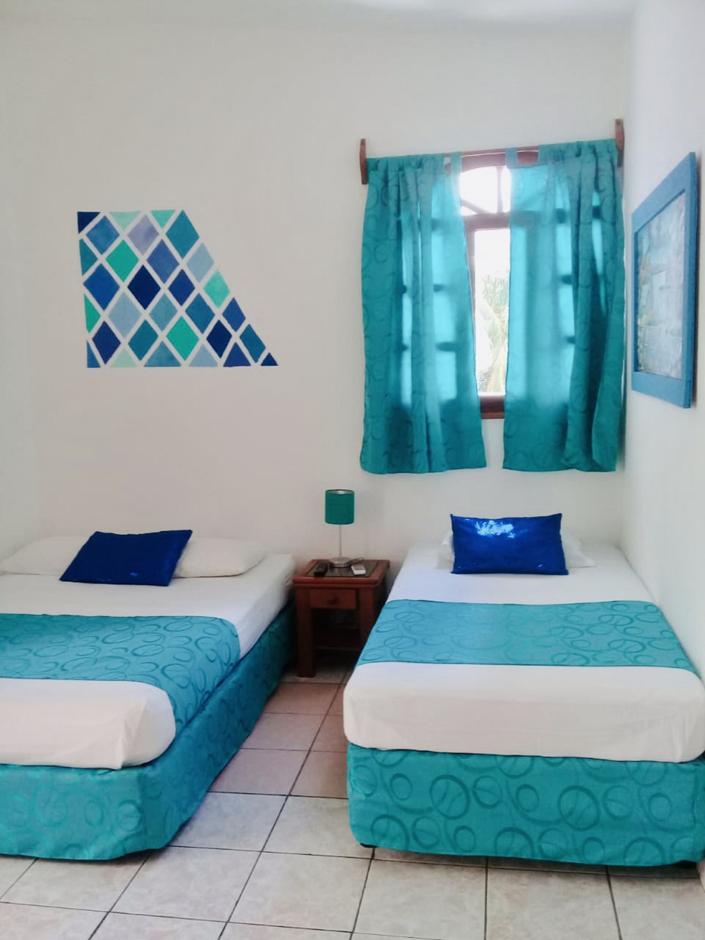 Une chambre avec deux lits, chacun recouvert d’une couvre-lit bleue et blanche, une petite table de chevet avec une lampe et une fenêtre ornée de rideaux turquoise.