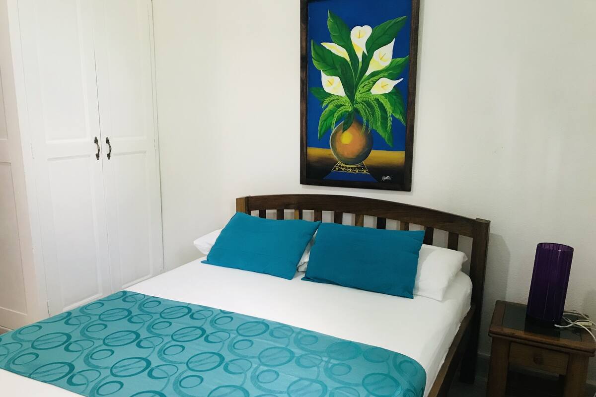 Een bed met blauw-witte beddengoed, een houten hoofdbord en een klein nachtkastje met een paarse vaas.