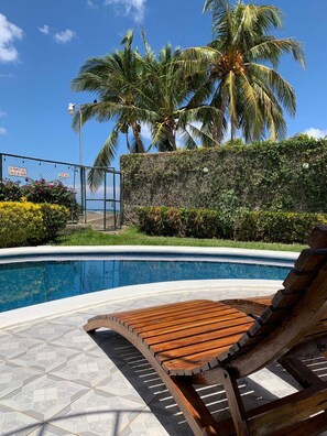 A swimming pool with a wooden lounge chair, surrounded by palm trees and a stone wall. - HC Liri Hotel (San Juan del Sur)