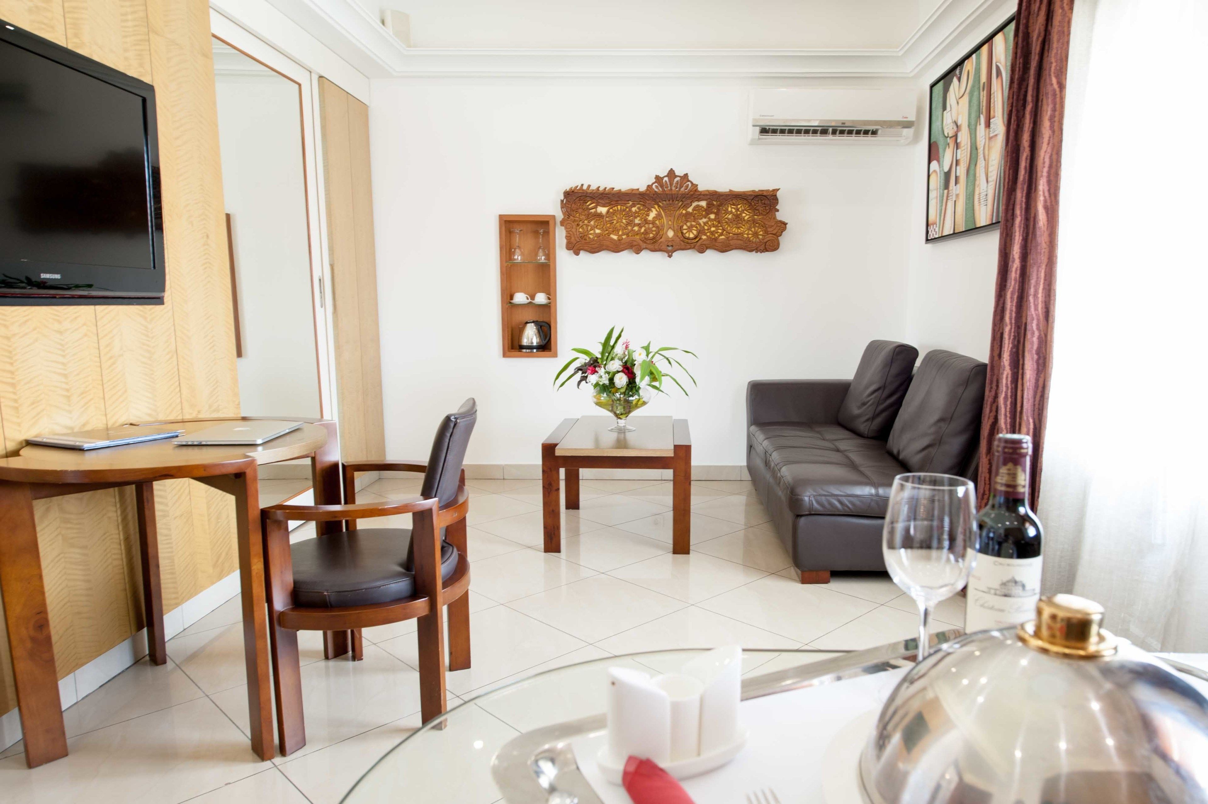 A modern living room with a flat-screen TV, wooden furniture, a sofa, a dining table with a vase of flowers, and a wine bottle.