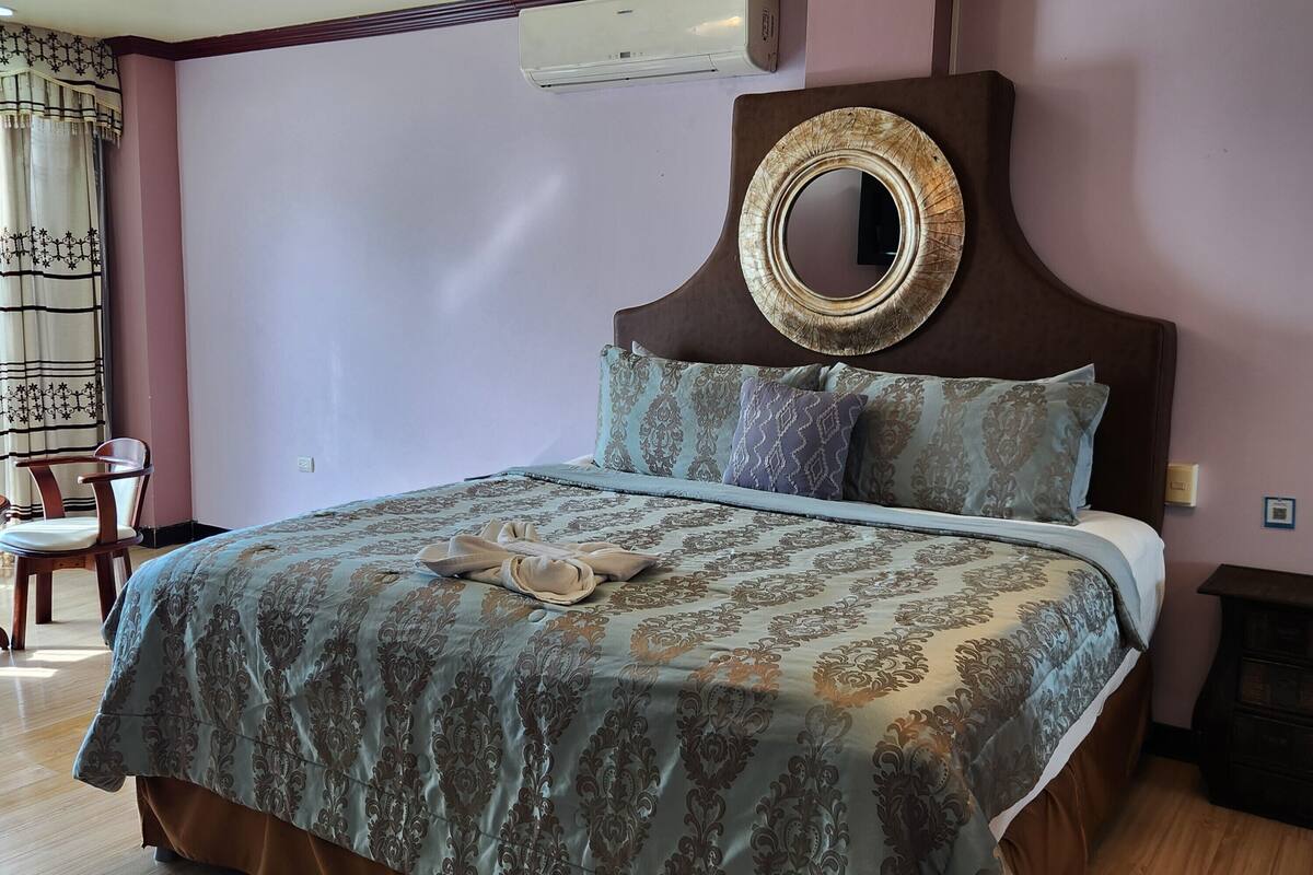 A bedroom with a bed, a wooden floor, a wall-mounted air conditioner, and a chair.