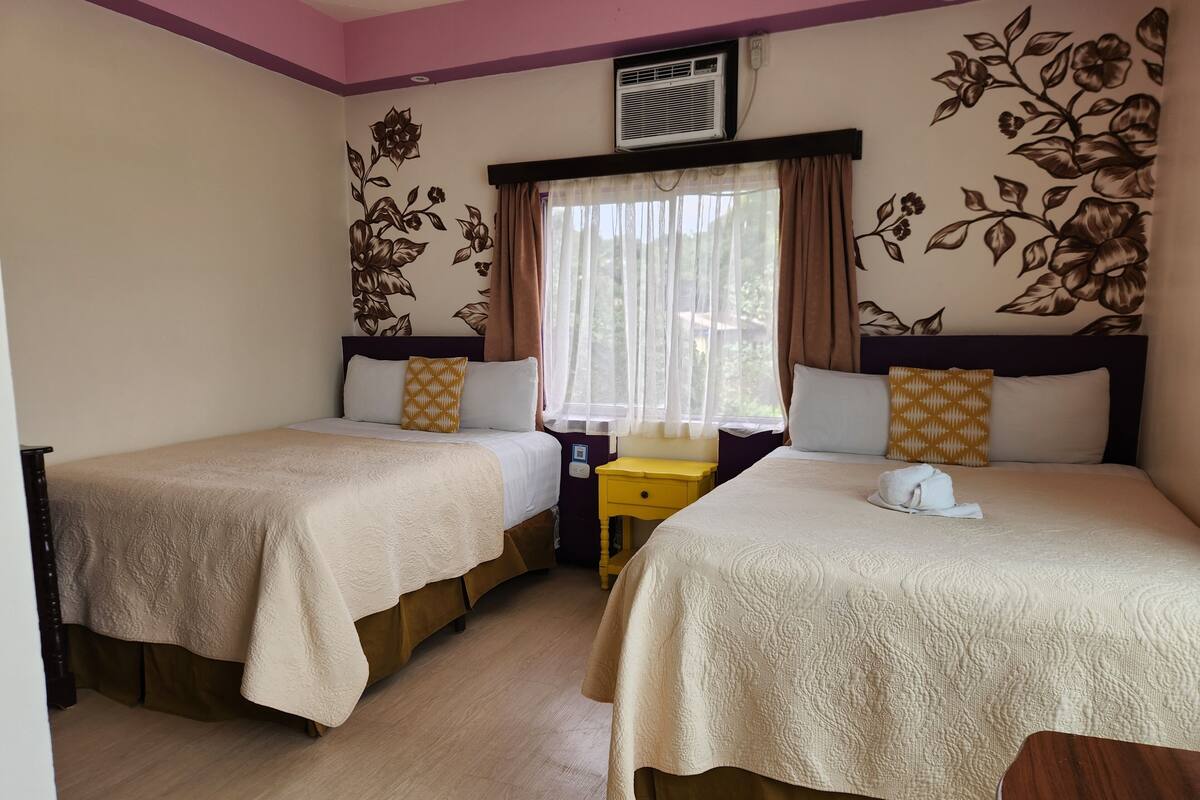 A hotel room with two beds, a window with sheer curtains, a wooden nightstand, and floral wall decorations.