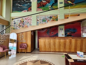 A hotel lobby with a staircase, wooden reception desk, and colorful murals on the walls.