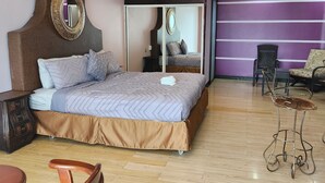 A bedroom with a bed, a mirror, a chair, a small table, and a wall with a purple stripe. - Park Place Hotel (Siguatepeque)