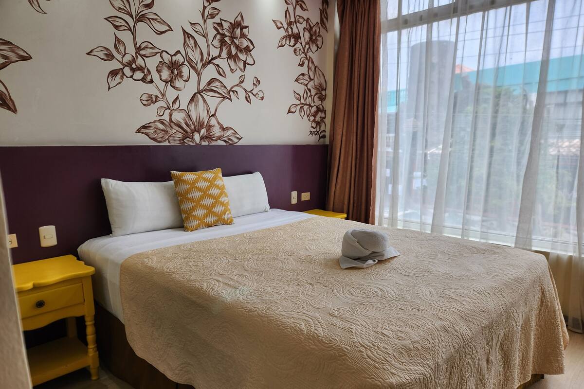 A hotel room with a bed, a nightstand, a yellow drawer, a patterned wall, and a window with curtains.