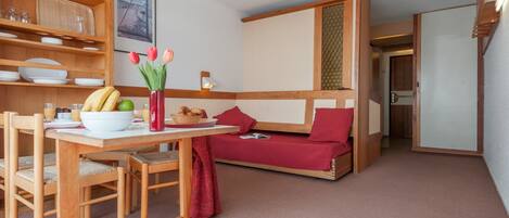 A cozy room with a wooden dining table, chairs, a red sofa, and a small kitchen area with shelves and a vase of flowers.