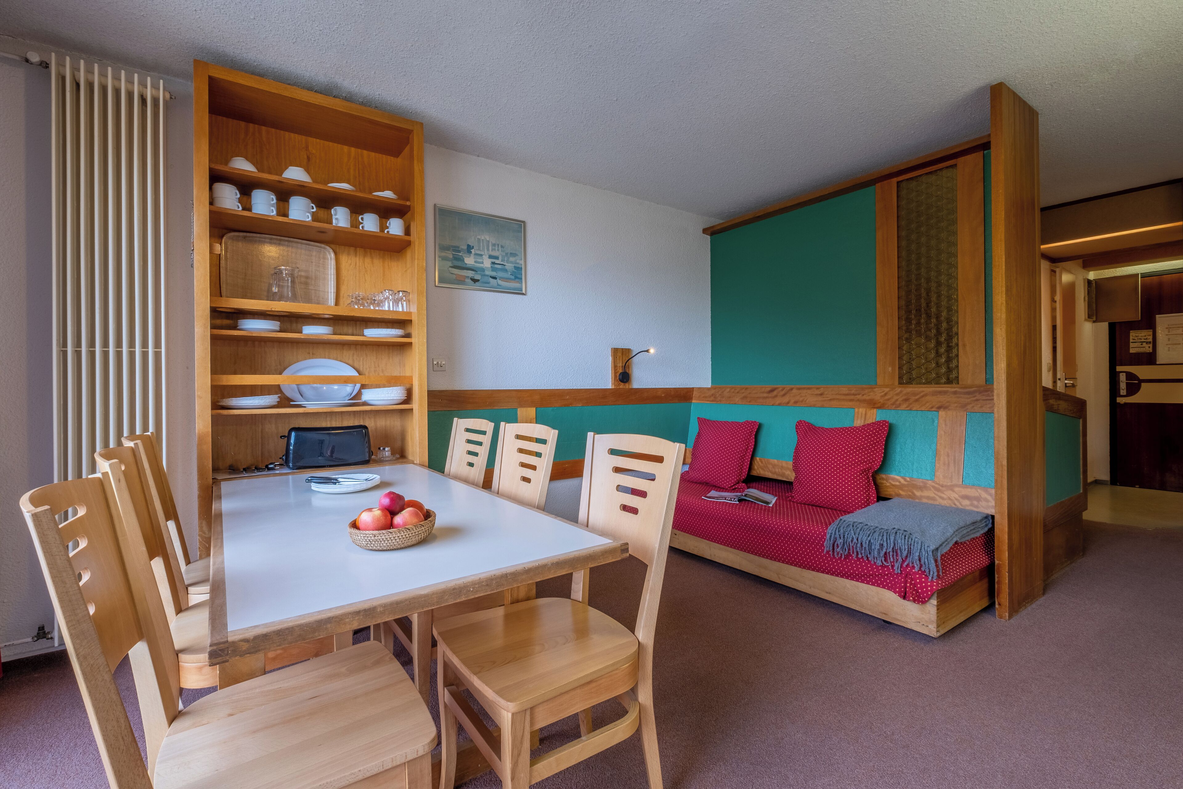 A dining area with a wooden table and chairs, a wooden cabinet with dishes, and a sofa with red cushions.