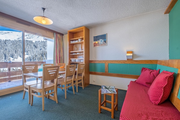 A cozy room with a wooden dining table and chairs, a red sofa, and a bookshelf. - Méribel Résidence Pierre & Vacances Les Bleuets (Les Allues)
