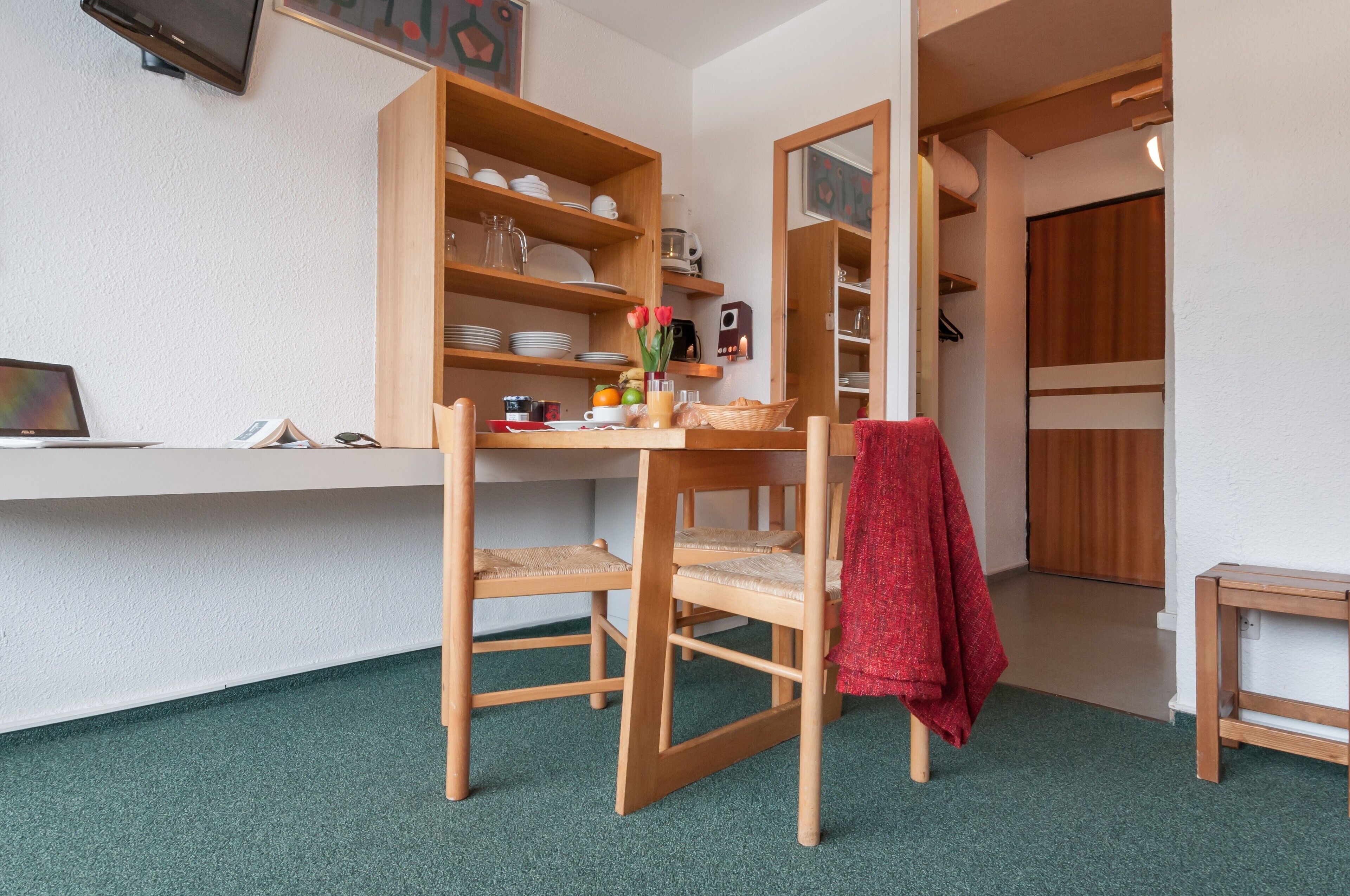 A compact room with a wooden desk, two chairs, a small table, and a shelf with dishes and a basket.