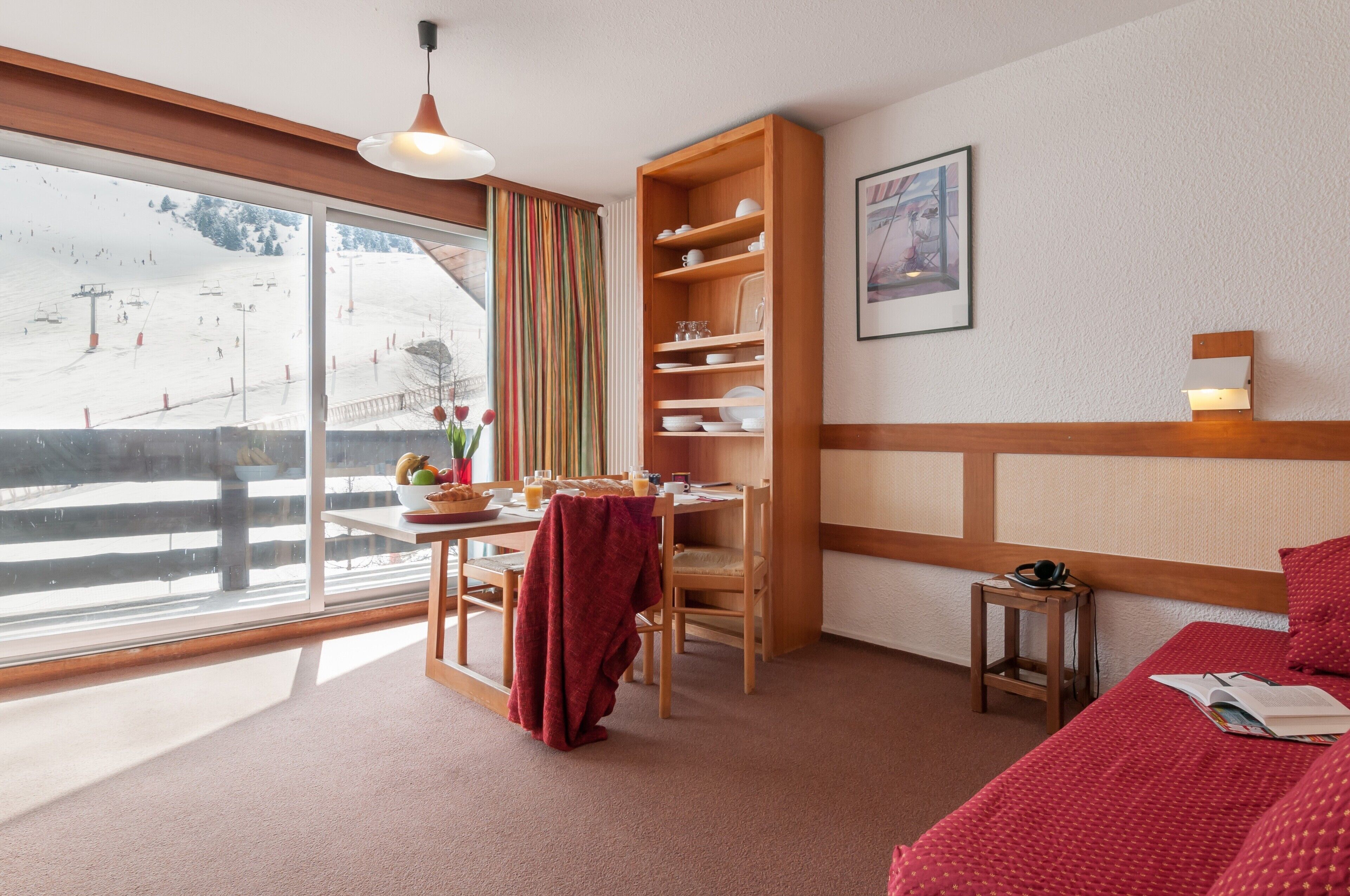Een gezellige kamer met een houten tafel, stoelen, een boekenplank, een bed met rode beddengoed en een groot raam met uitzicht op een besneeuwd landschap.