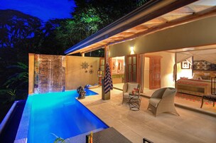 Un área de piscina con una piscina azul cristalina, un muro de piedra y una pérgola de madera. Hay sillas de mimbre y una pequeña mesa con una bebida.