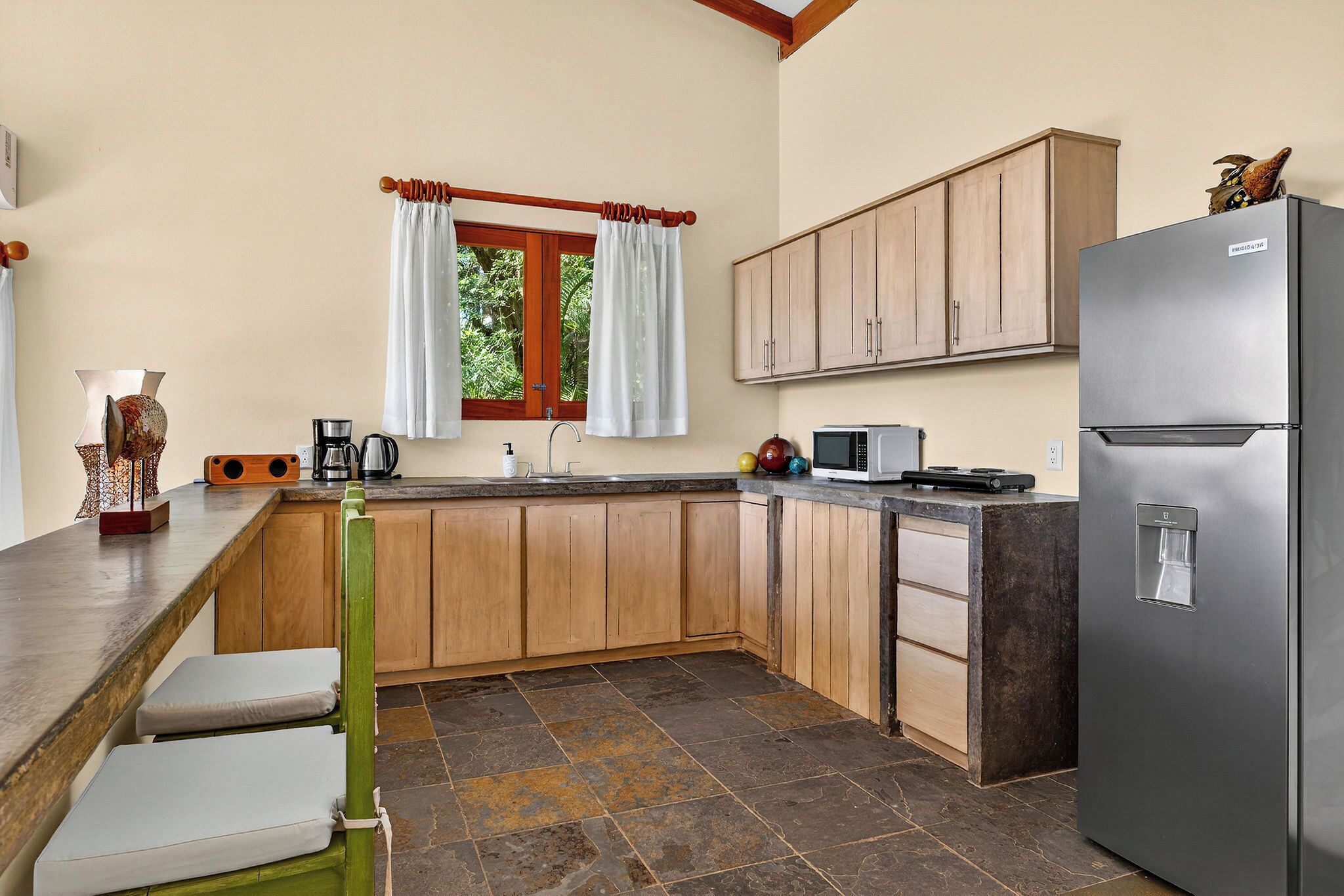 A kitchen with wooden cabinets, a stainless steel refrigerator, a window with curtains, and a stone-tiled floor.