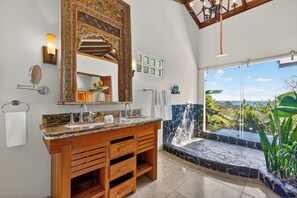 Un baño con un espejo grande, dos lavamanos, un mueble de tocador con cajones, un perchero y vistas a un paisaje montañoso a través de un ventanal amplio.