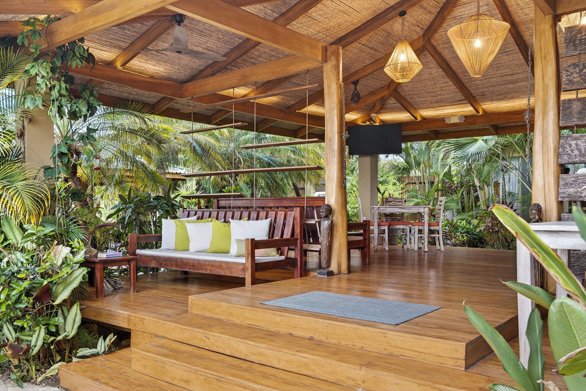 Um deck de madeira com um sofá e cadeiras, rodeado por plantas tropicais exuberantes.