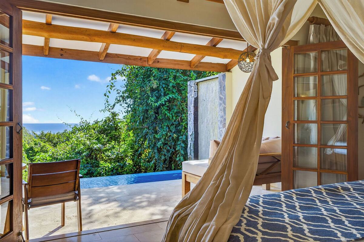 Ein Schlafzimmer mit Holzbalkendecke, einem Bett mit gemustertem Überwurf, einem Stuhl und Blick auf Grünflächen sowie einen Pool.