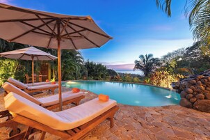 Un área junto a la piscina con sillones y sombrillas, rodeada de vegetación tropical y un cielo despejado.