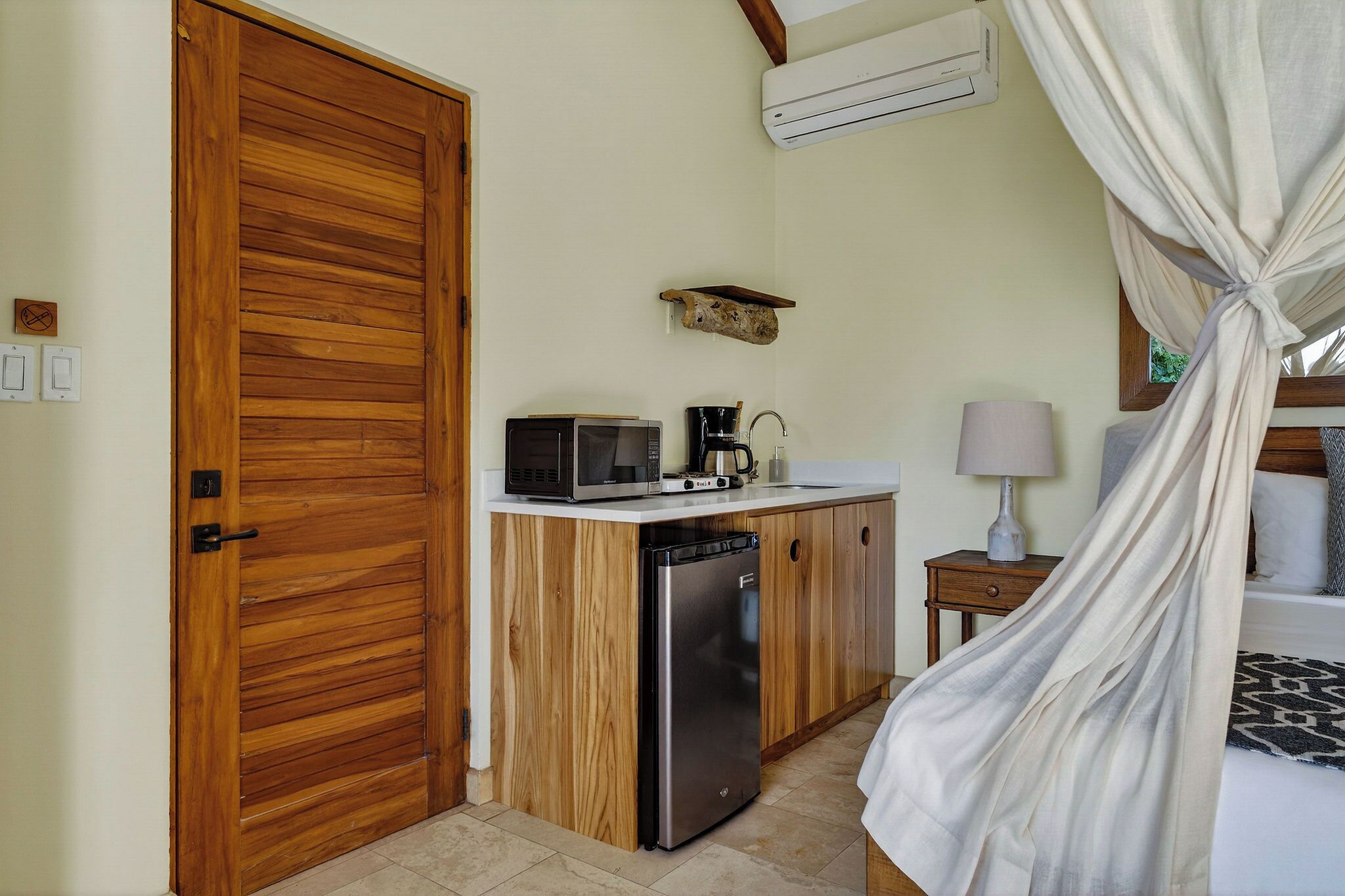 Une chambre de taille modeste, dotée d’une porte en bois, d’un four à micro-ondes, d’une machine à café, d’un petit lavabo, d’une table de chevet avec une lampe et d’un lit à baldaquin.