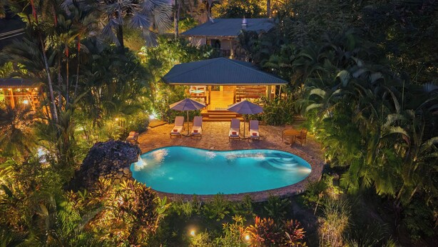Un complexe hôtelier doté d’une piscine, de chaises longues et d’un espace salon abrité, le tout entouré d’une végétation luxuriante.