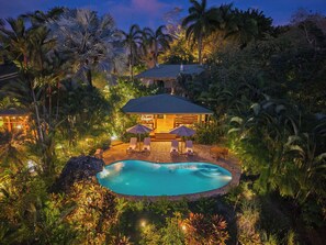 Un resort con piscina, sillas de descanso y un área de estar techada, rodeada de exuberante vegetación.