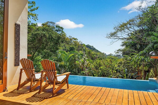 Eine Holzterrasse mit zwei Adirondack-Stühlen, einem Swimmingpool und üppigem Grün im Hintergrund.