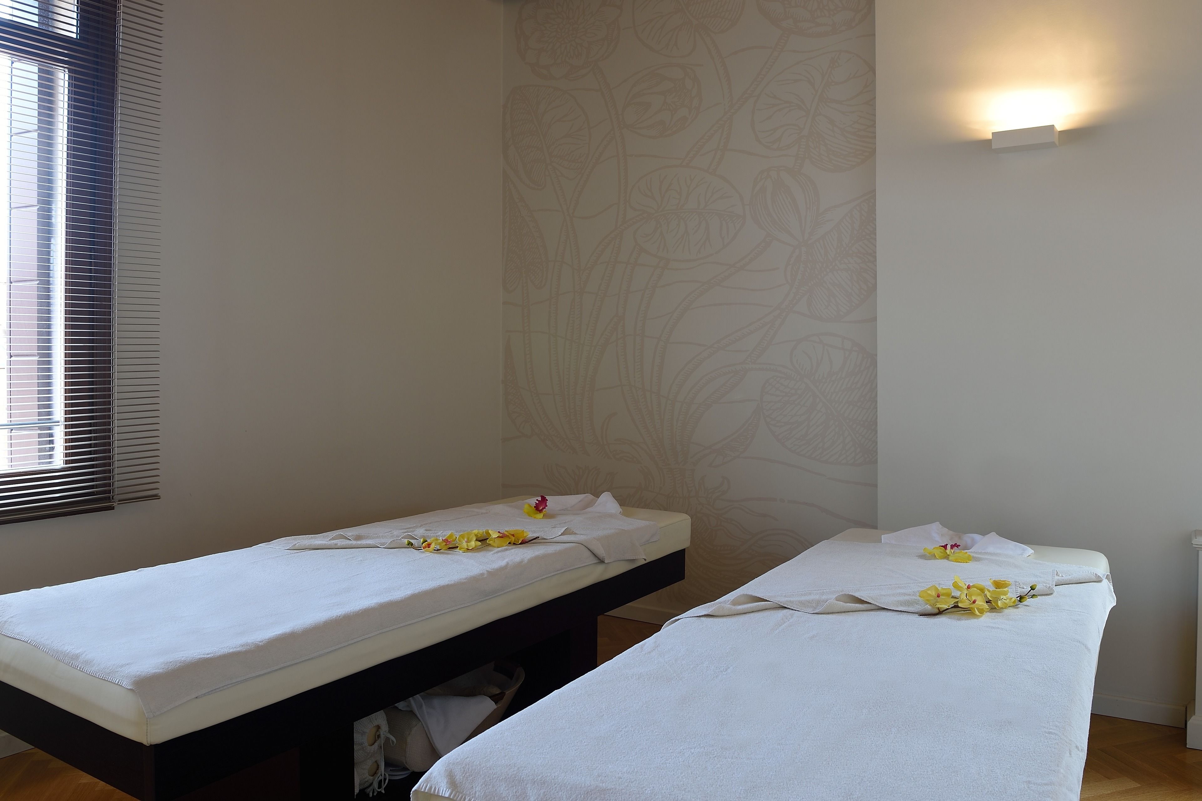 Dos camillas de masaje con sábanas blancas y flores amarillas, una lámpara fijada a la pared y una ventana con persianas.