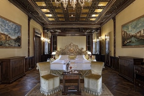 A luxurious hotel room with a large bed, ornate ceiling, and classic paintings.