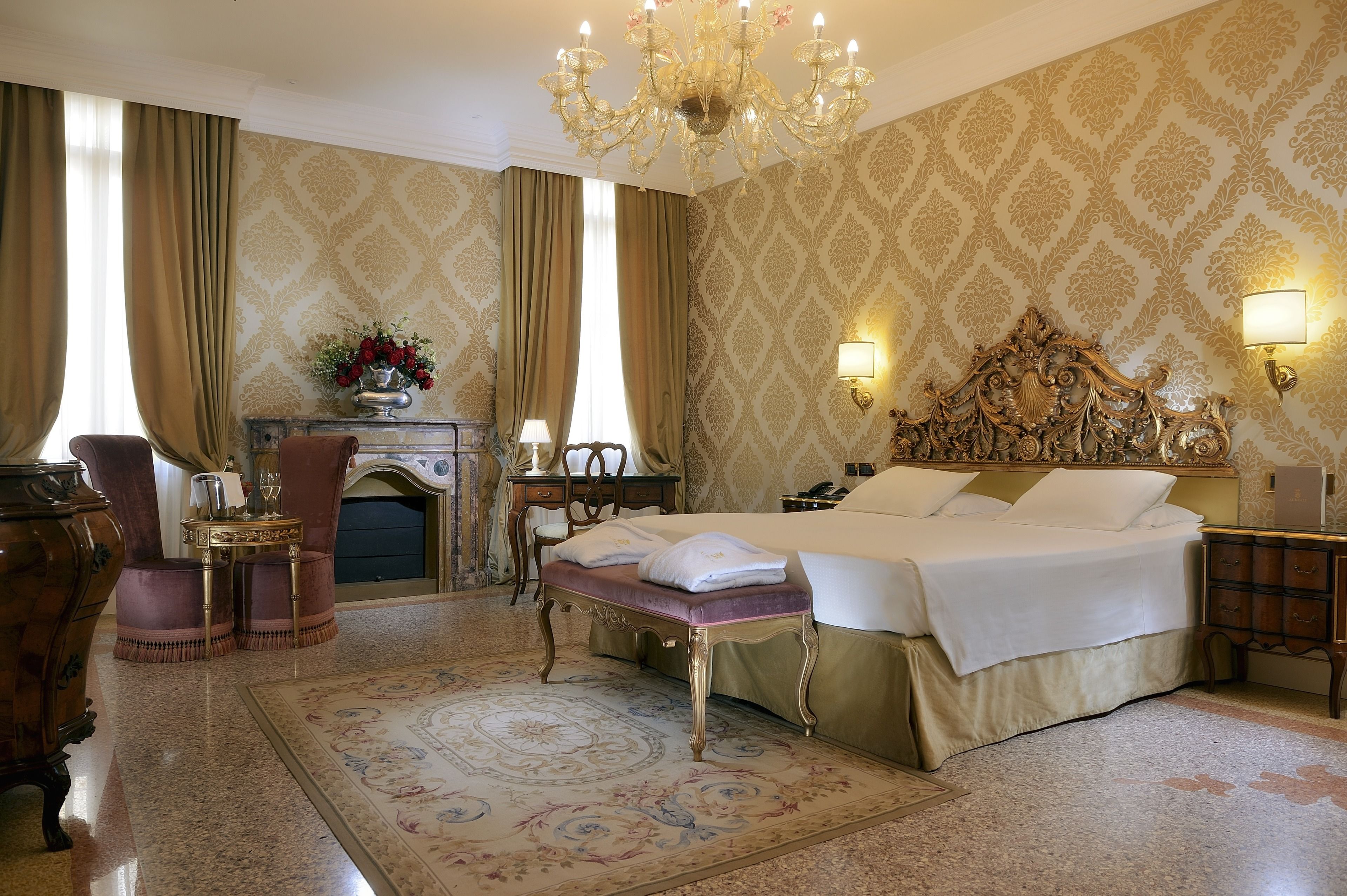 Quarto de hotel com uma cama grande, um candelabro, uma escrivaninha com abajur e uma lareira.