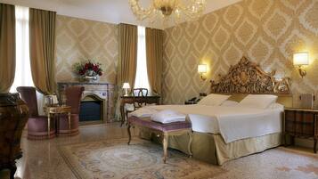 Quarto de hotel com uma cama grande, um candelabro, uma escrivaninha com abajur e uma lareira.