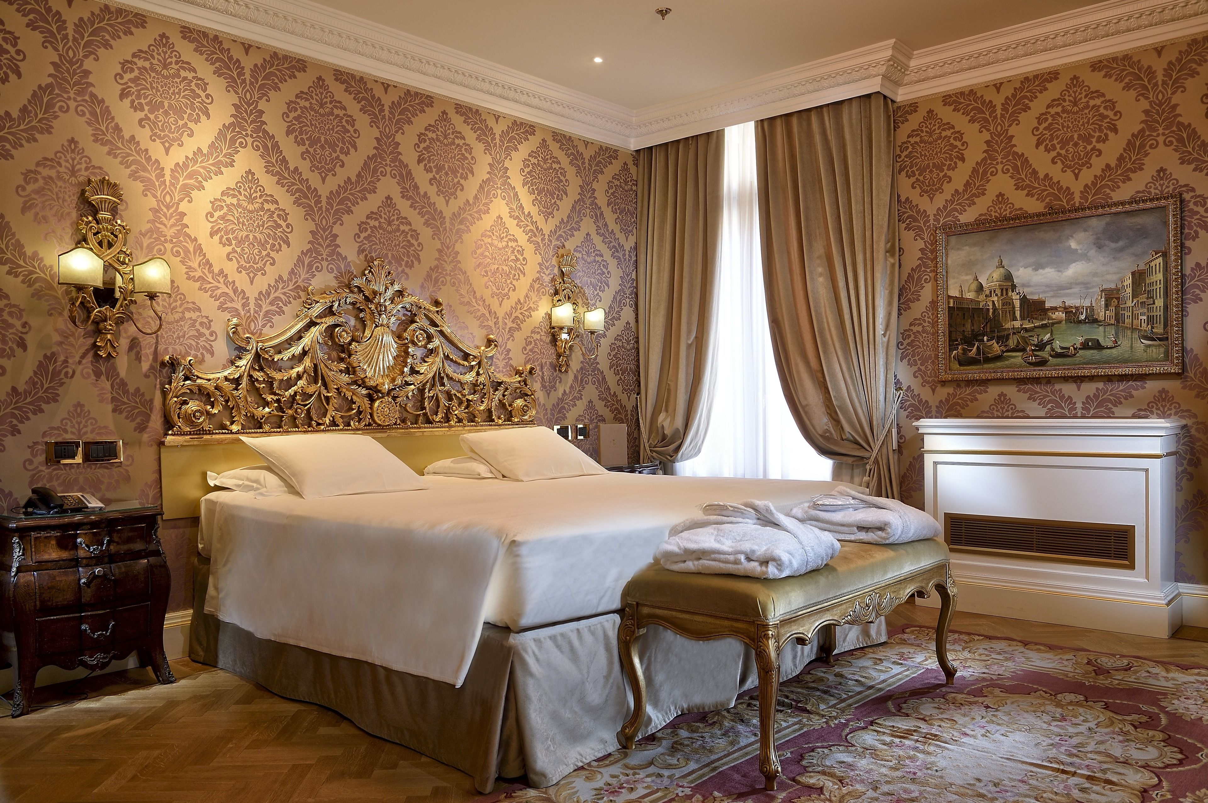 A luxurious bedroom with a large bed, ornate headboard, wall sconces, a fireplace, and a classic painting.