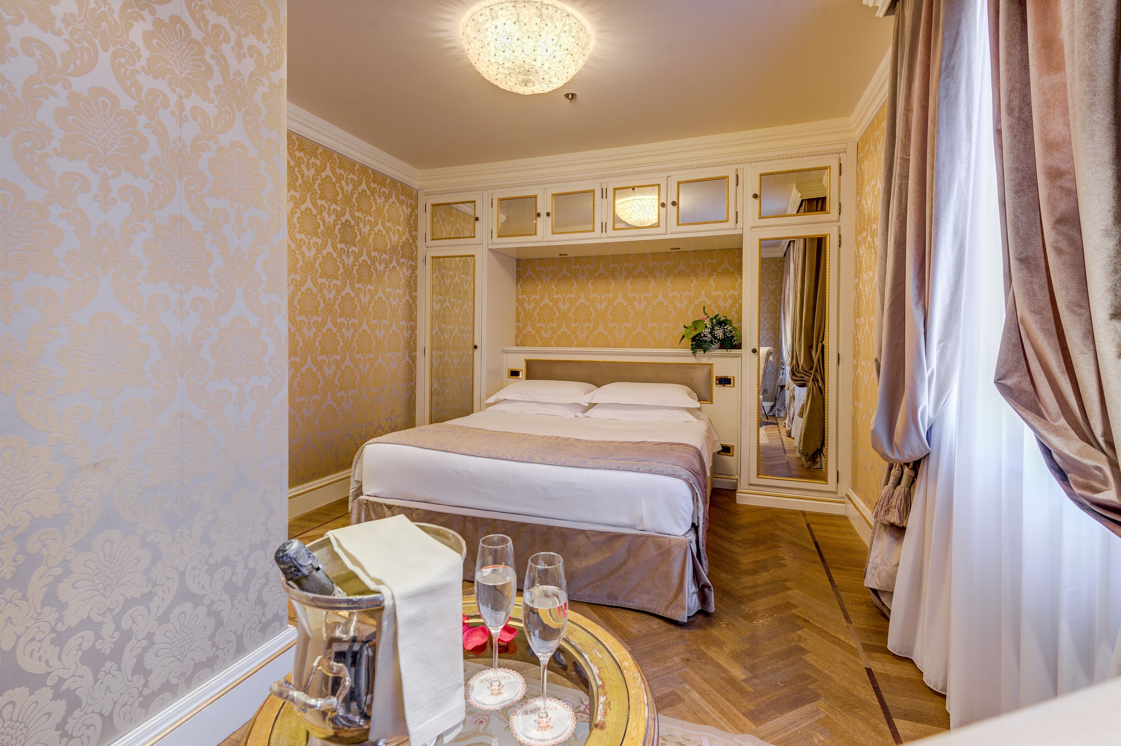 A hotel room with a large bed, a mirror above the bed, a round table with a tray, and a chandelier.