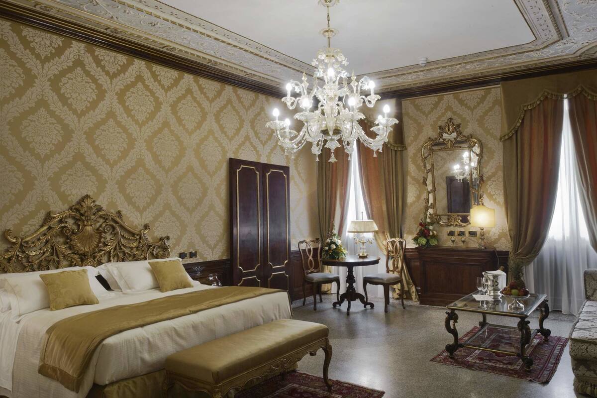 A luxurious bedroom with a large bed, a chandelier, a sitting area with a table, and ornate decor.