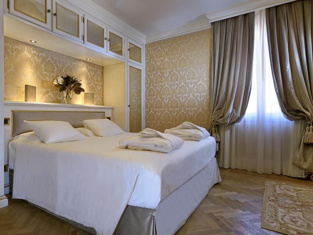 Ein Hotelzimmer mit einem großen Bett, einem Spiegel darüber, einer Vase mit Blumen und einem Fenster mit Vorhängen.