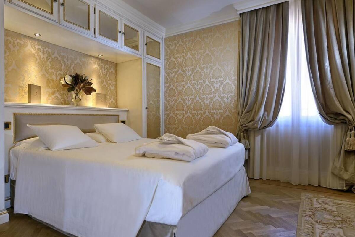 A hotel room with a large bed, a mirror above it, a vase with flowers, and a window with curtains.
