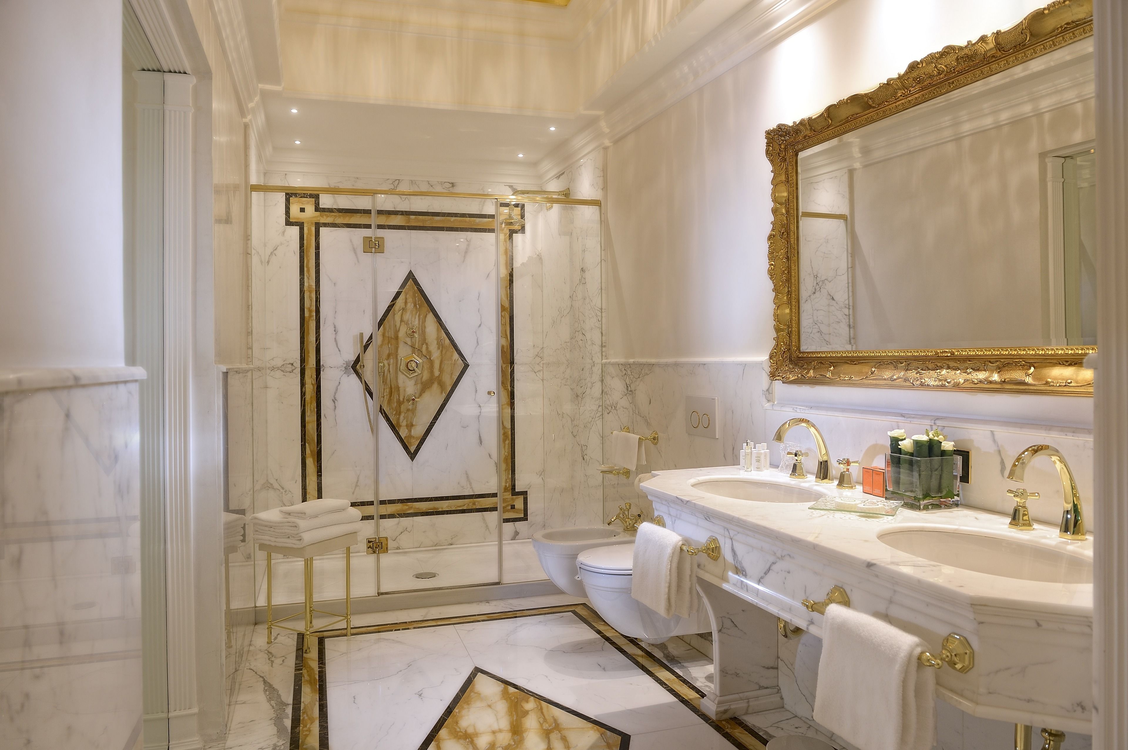 Un baño lujoso con paredes de mármol, un espejo con marco dorado, lavamanos dobles y cabina de ducha.