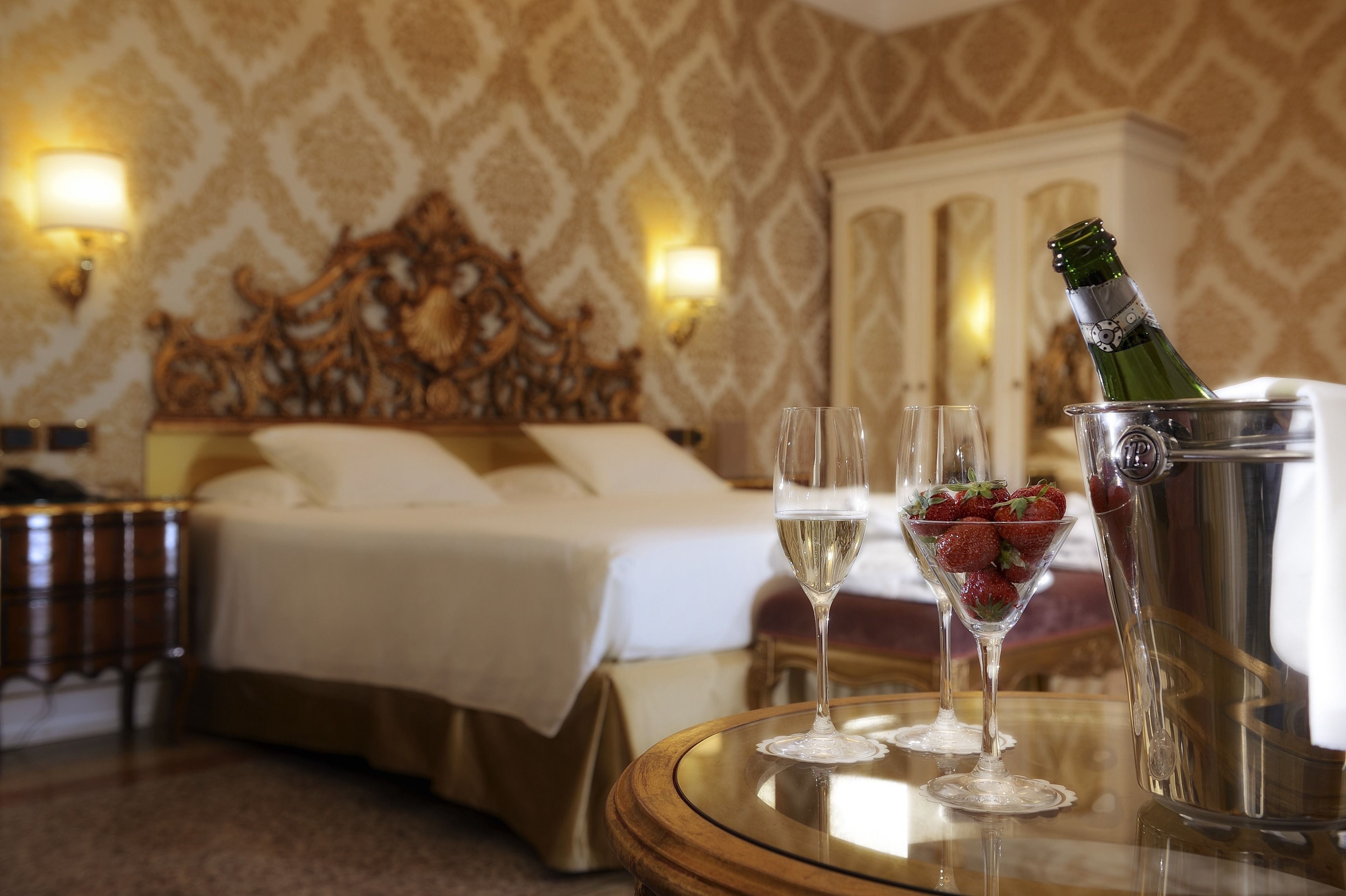 A hotel room with a bed, a bedside table with a lamp, a minibar, and a tray table with a bottle of champagne and two glasses.