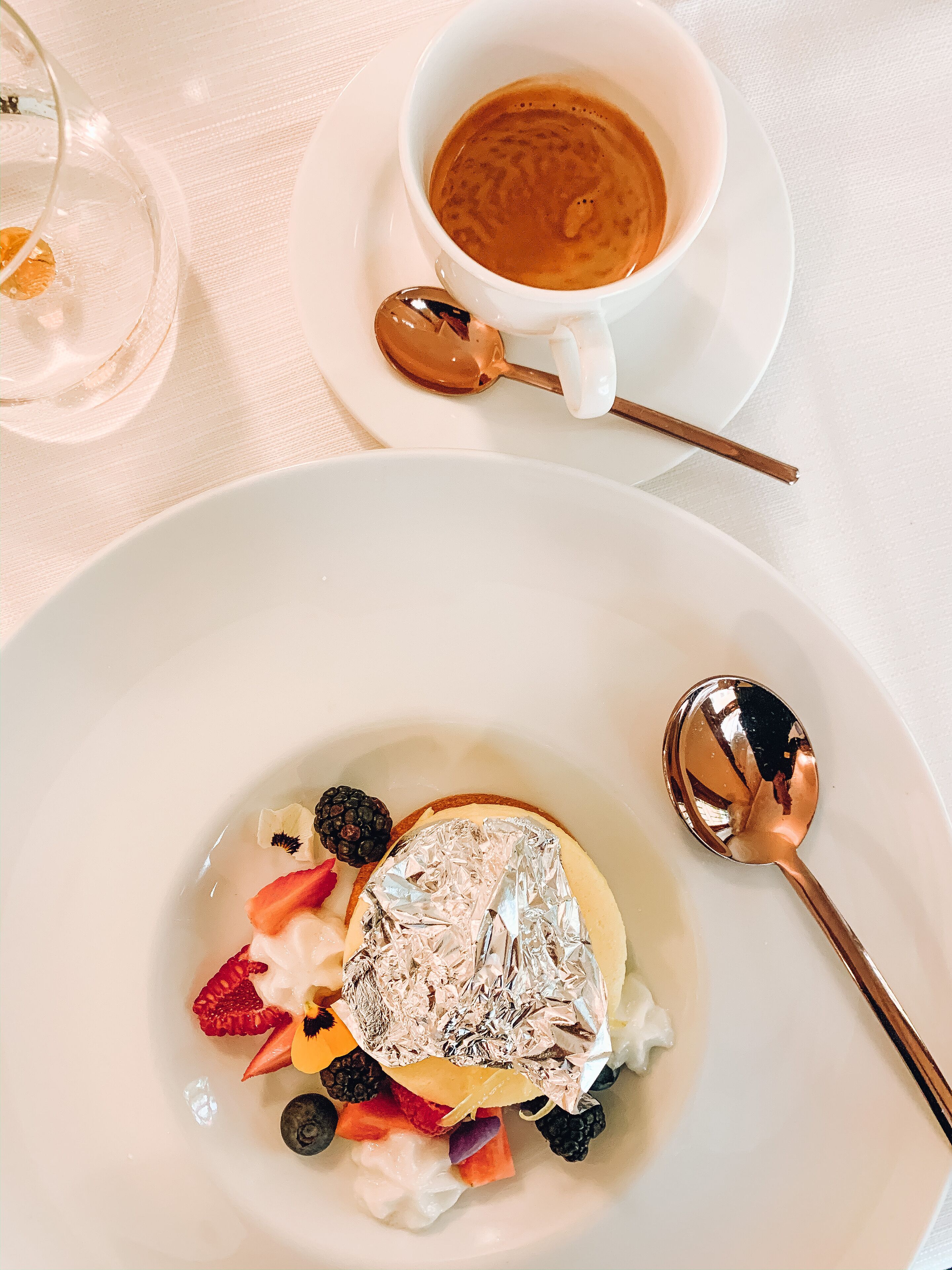 A dessert plate with fruit and a foil-wrapped item, a cup of coffee, and a spoon on a white tablecloth.