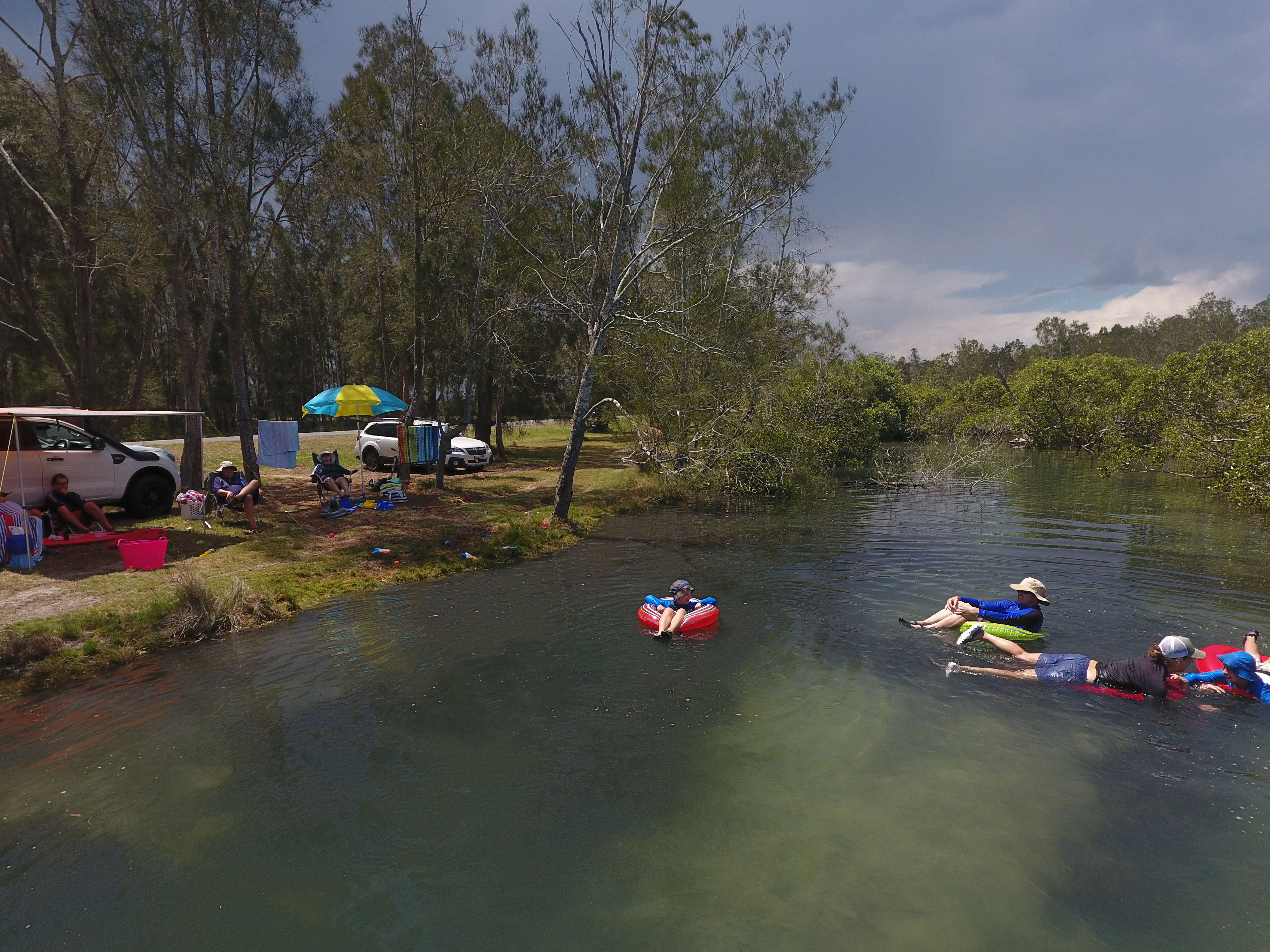 Une rive de rivière où des personnes se détendent, et un véhicule est garé à proximité.