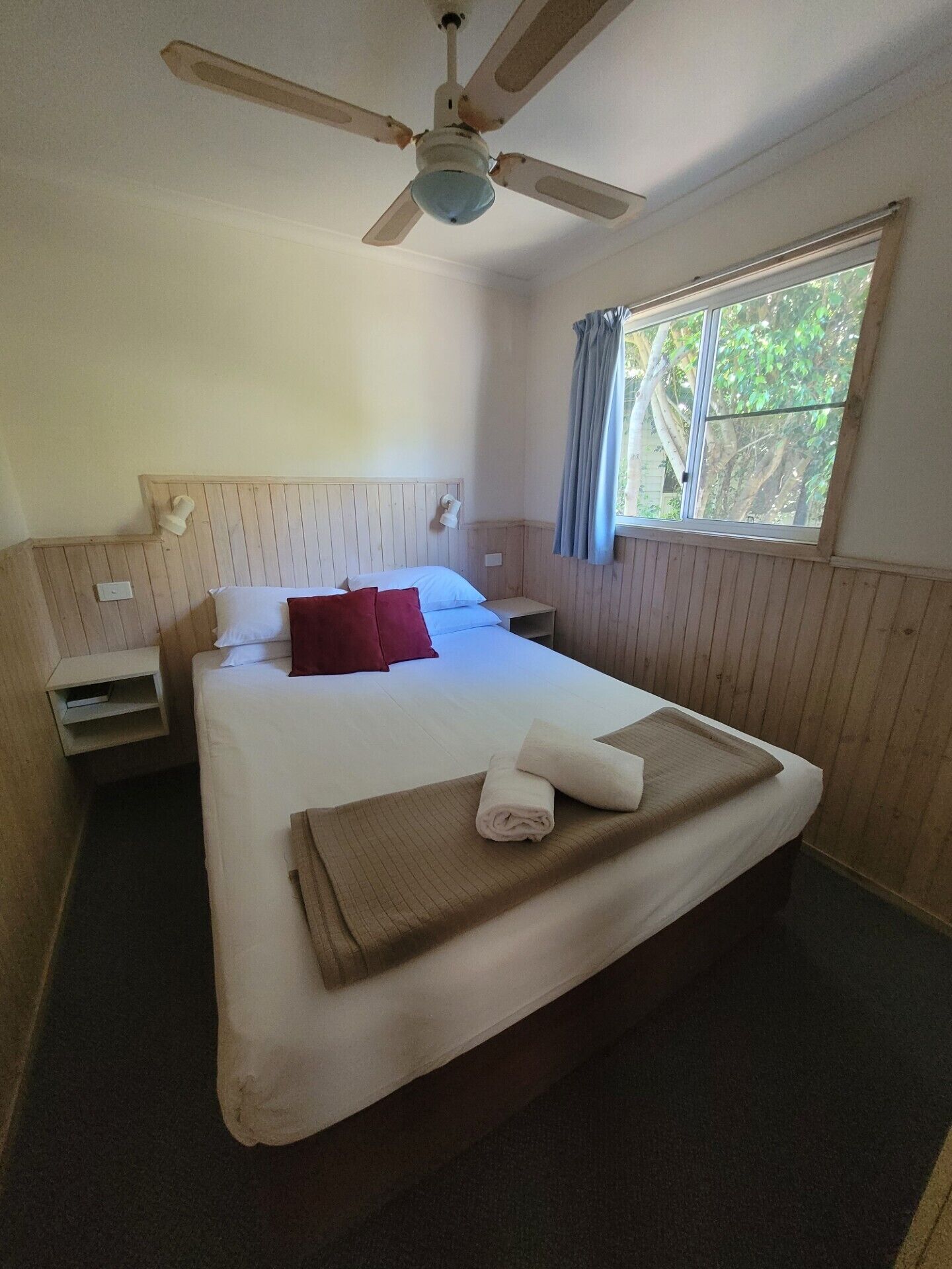 Habitación de hotel con una cama grande, un ventilador de techo, una ventana con cortinas y una mesita de noche con lámpara.