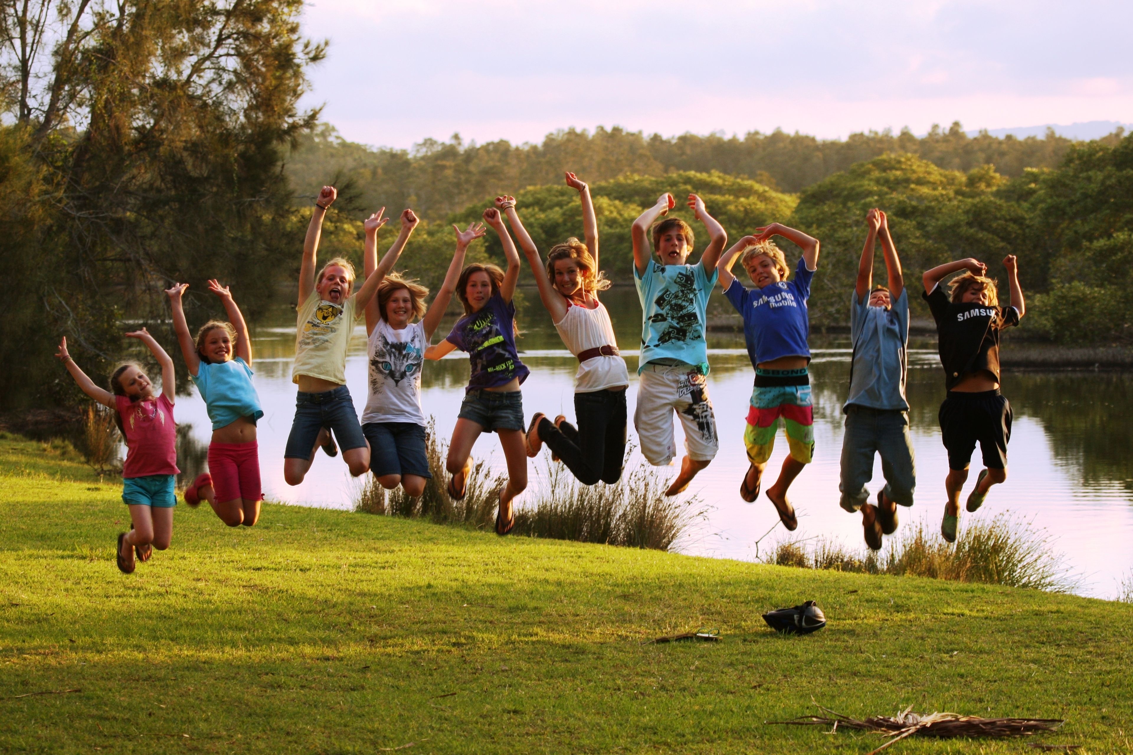 A group of people jumping in a grassy area near a body of water with trees in the background.