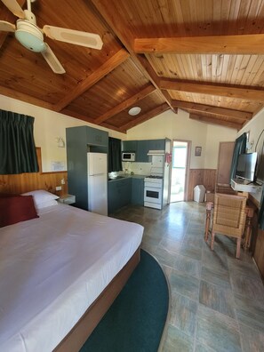 Un dormitorio con cama, ventilador de techo, kitchenette con microondas, estufa y lavaplatos, y baño con ducha e inodoro.