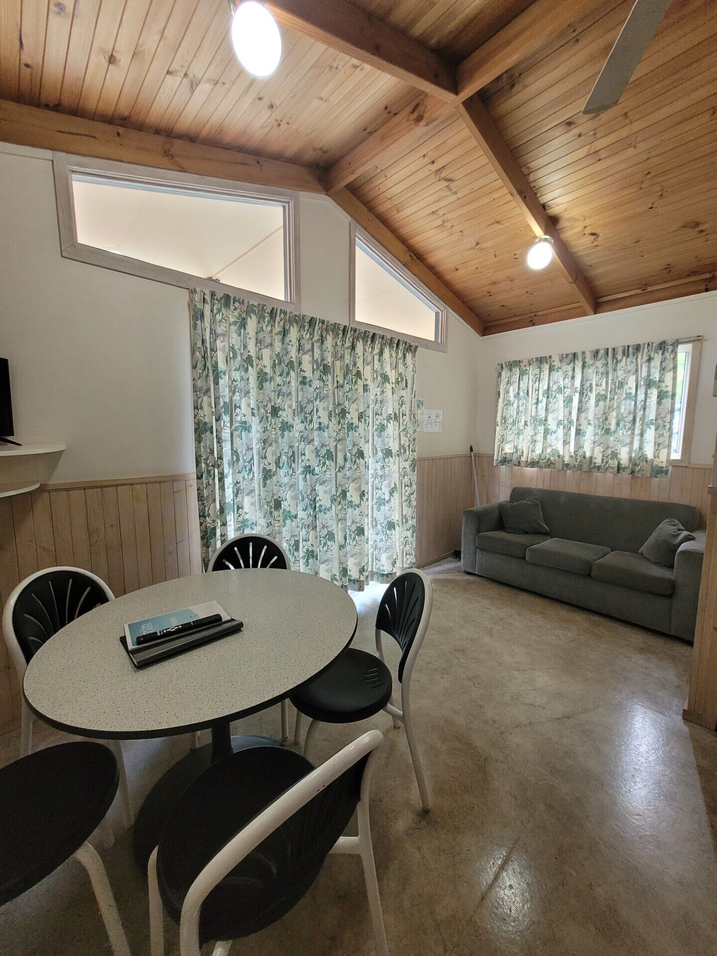 A cozy room with a wooden ceiling, a round table with chairs, a sofa, and floral curtains.