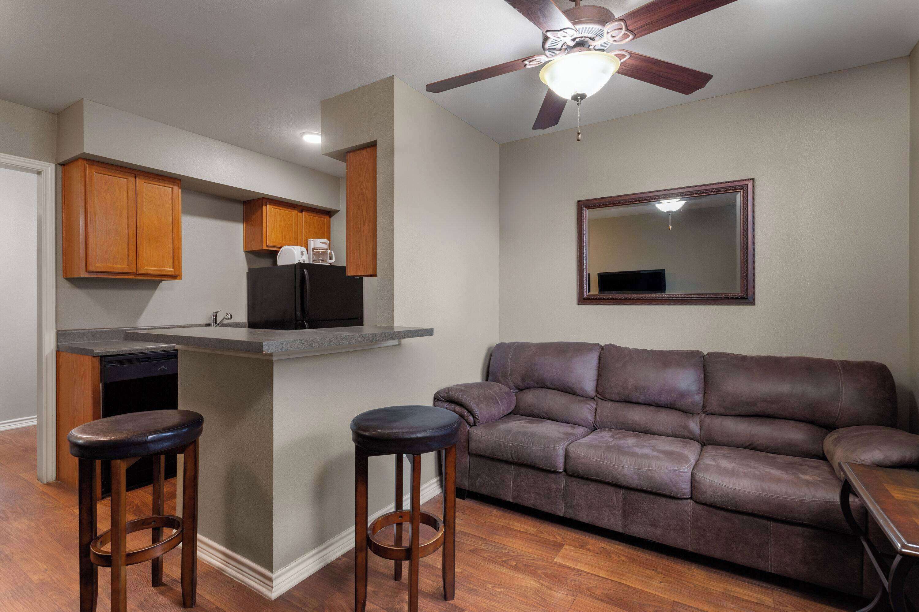 A compact living space with a kitchenette, a brown leather sofa, two wooden bar stools, and a ceiling fan.