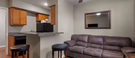 A compact living space with a kitchenette, a brown leather sofa, two wooden bar stools, and a ceiling fan.