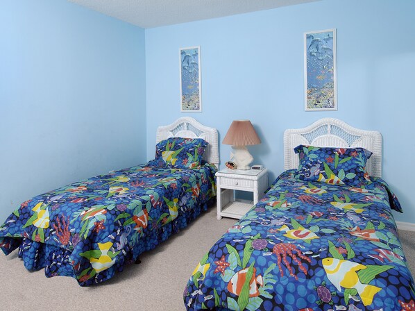 A bedroom with two single beds, each with a white headboard and a colorful bedspread featuring marine life. There is a nightstand with a lamp and a decorative fish between the beds.