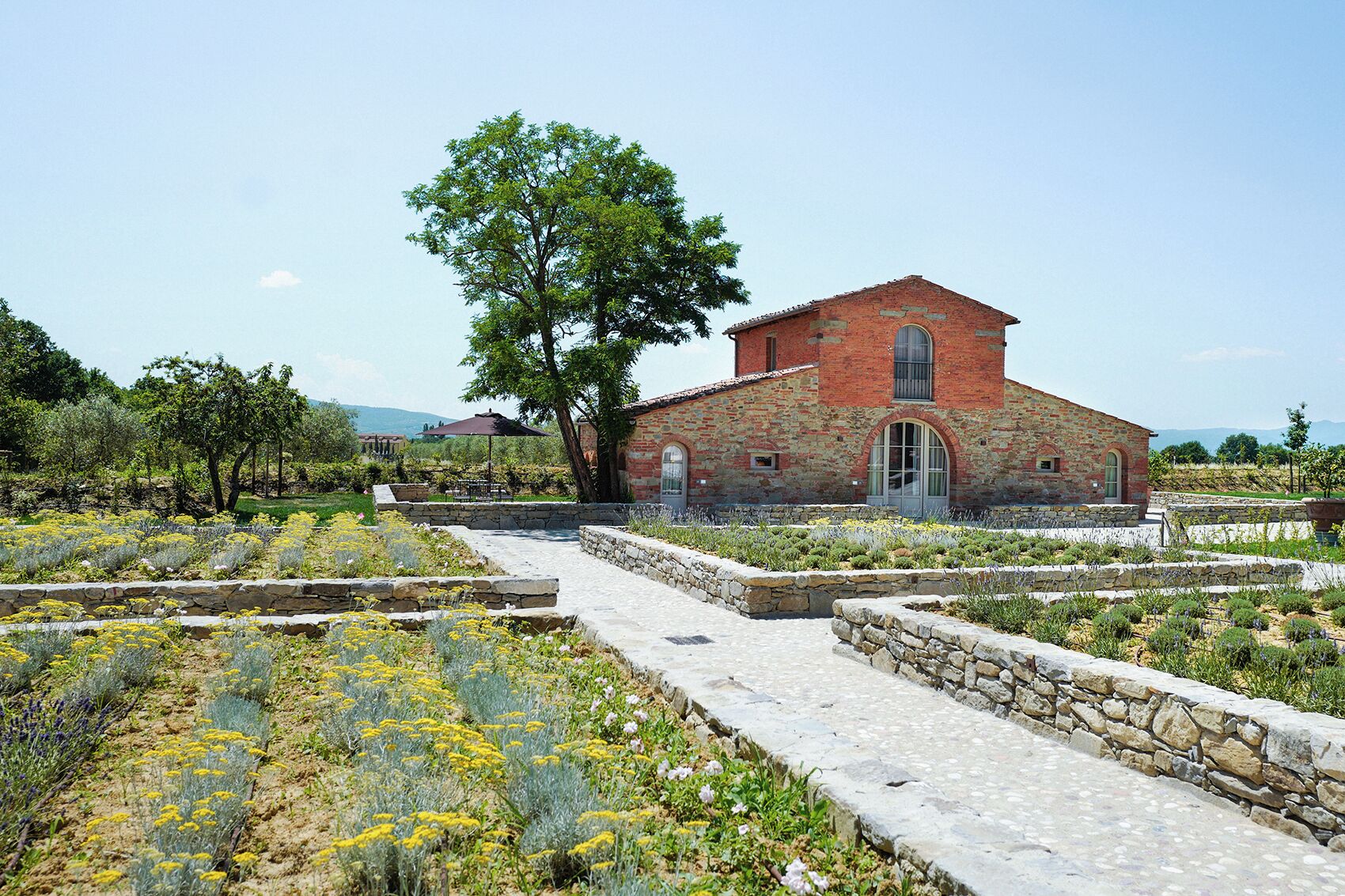 A red brick building with a stone pathway leading to it, surrounded by a garden with various plants and flowers.