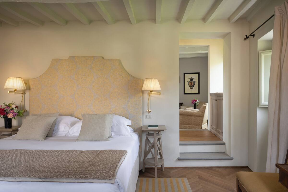 Un dormitorio con una cama grande, dos lámparas de noche, una mesita de noche con un jarrón de flores y vistas a un pasillo que conduce a un baño.