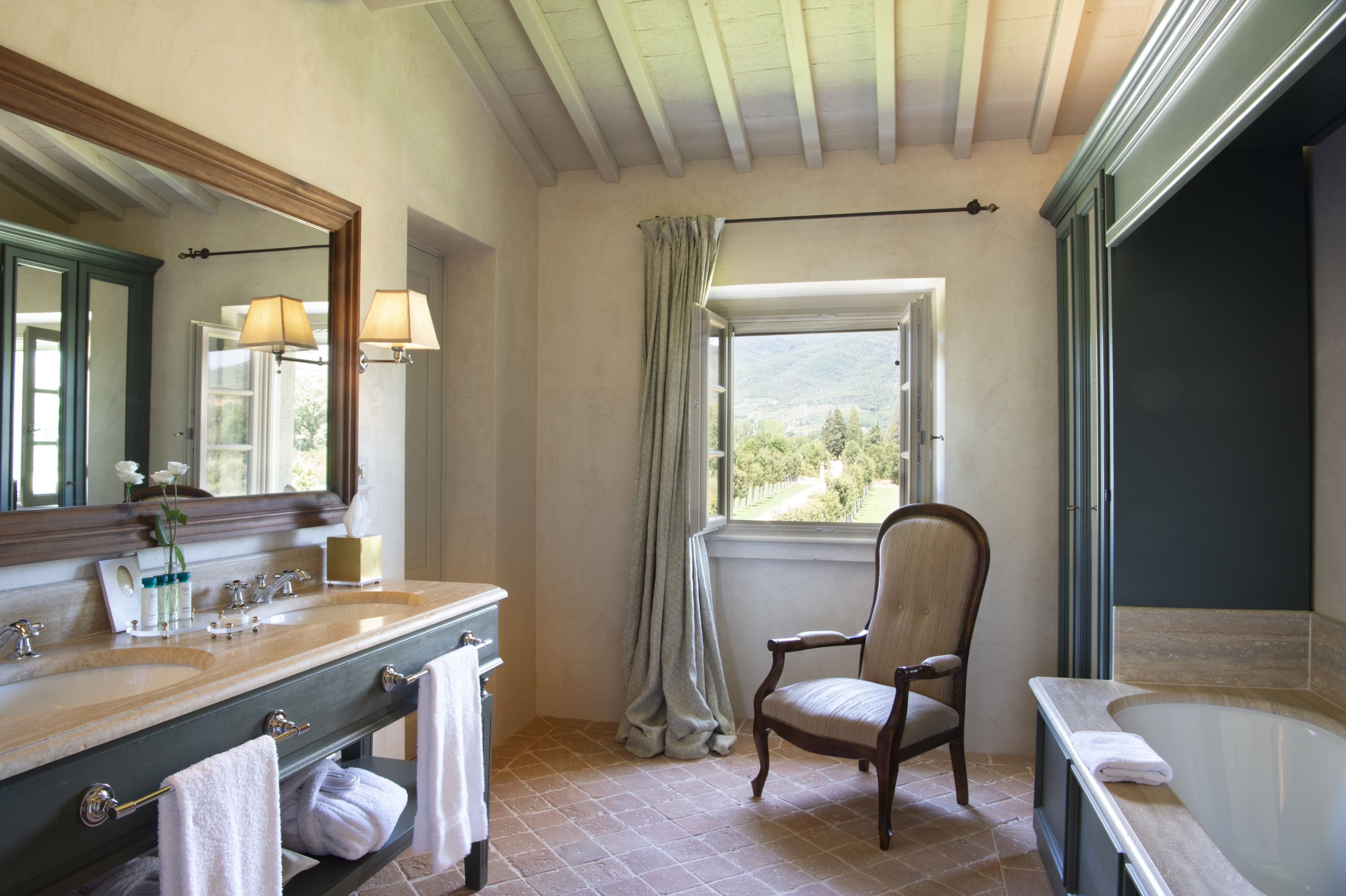 A bathroom with a large mirror, two sinks, a chair, a window with curtains, and a bathtub.