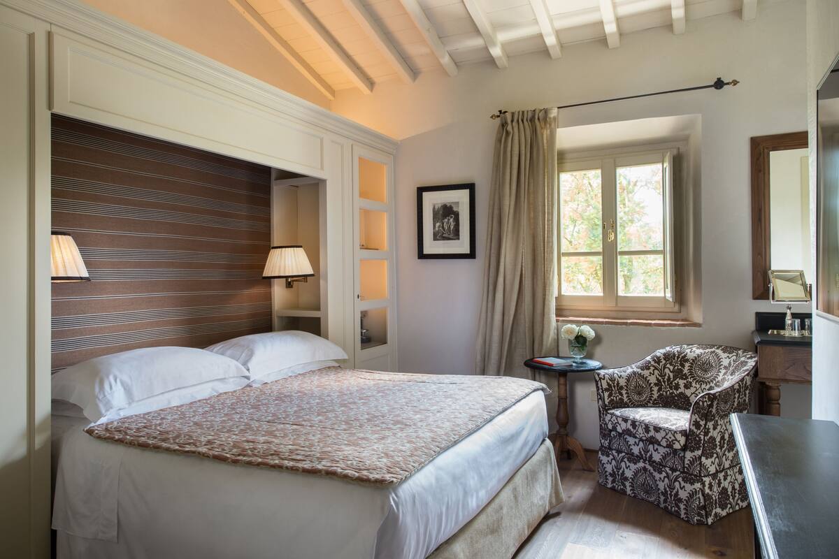 A bedroom with a bed, bedside lamps, a chair, a small table, and a window with curtains.